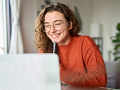 Young woman in glasses and an orange sweater smiling at a laptop screen