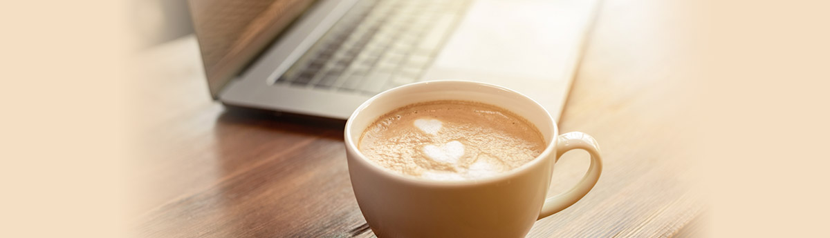 A latte in a beige mug with a heart design in the foam next to a laptop 