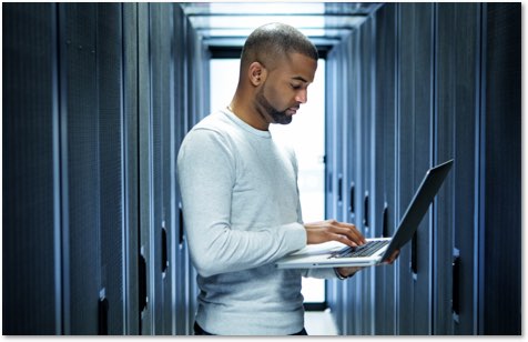 black man in a white sweater standing in a server room holding a laptop