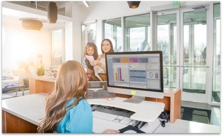 Woman behind a reception desk greeting a patient coming in with a child in her arms