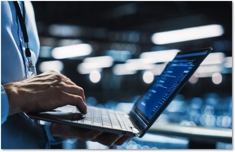 Man in a dress shirt and lanyard holding a laptop and typing on it