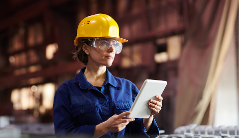 Woman in a hardhat and safety glasses holding a tablet computer on a factory floor
