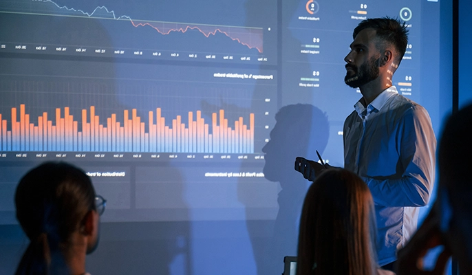 Man presenting a graph on a wall screen in a darkened conference room
