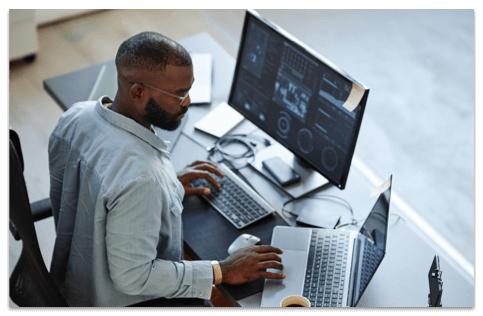 Bald black man with a beard working on a desktop and a laptop computer at the same time