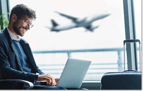 Man with short, curly brown hair and glasses smiling at a laptop at an airport gate with a plane departing behind him