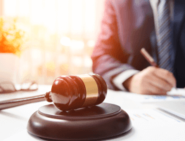 Foto: Person, die hinter einem weißen Schreibtisch schreibt, auf dem ein dunkler Auktionshammer liegt