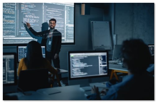 Teacher pointing at a screen in front of students working on monitors with code on the screens