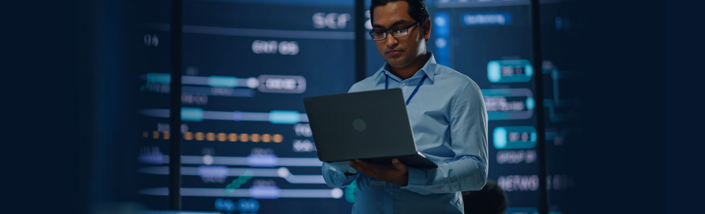 Man in glasses holding an open laptop in front of a large server room diagram