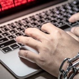 Hands chained with a padlock typing on a laptop showing a ransomware attack warning; symbolizing data encryption and cyber extortion.