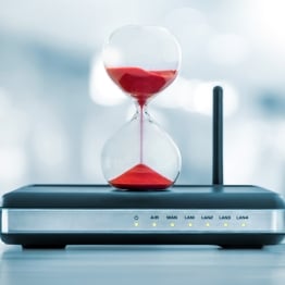 Hourglass sitting on a network router, symbolizing the shrinking time cybersecurity teams have to patch vulnerabilities before attackers exploit them.