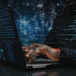 Person using a laptop surrounded by overlaid computer code, representing AI-powered cyberattacks, hacking, and cybersecurity threats.