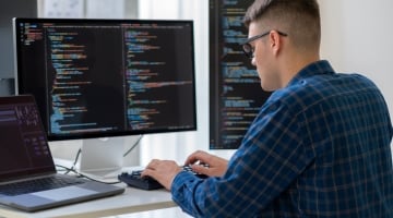 Developer working at a multi-monitor coding workstation with software code displayed across screens in a modern office environment.