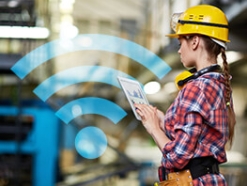 Woman in a hardhat working on a tablet computer in a factory with a blue wi-fi icon in the background