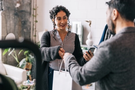 Woman with curly black hair handing a shopping bag to a man with a beard in a small business