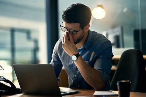 Man with short brown hair and glasses sitting at an office desk holding his head like he has a headache