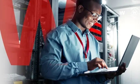 Smiling man working on a laptop he is holding up while in a server room