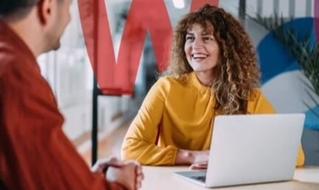 curly haired woman in a yellow top smiling at a meeting with her laptop open