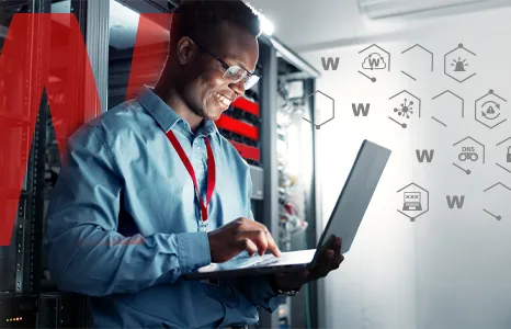 man in a blue shirt standing in a server room working on a laptop