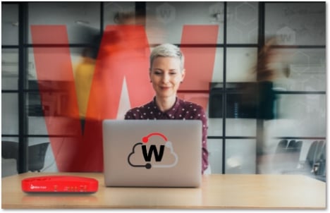 Woman with short white hair working at a laptop with a WatchGuard Cloud icon on the back and a red Firebox T115-W on the desk