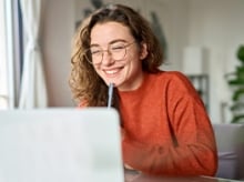 Young woman in glasses and an orange sweater smiling at a laptop screen