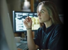 Blond woman working at a desk with a monitor behind her 