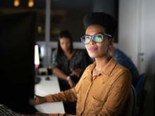 Mujer con gafas grandes y una camisa marrón de mangas largas trabajando frente a un monitor 