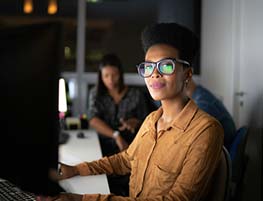 Frau mit großer Brille und braunem Langarmhemd arbeitet an einem Monitor 