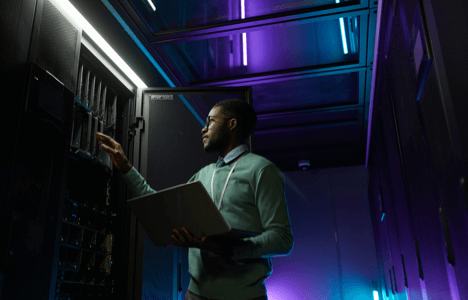 Man with a laptop in a dark server room lit with purple and green neon