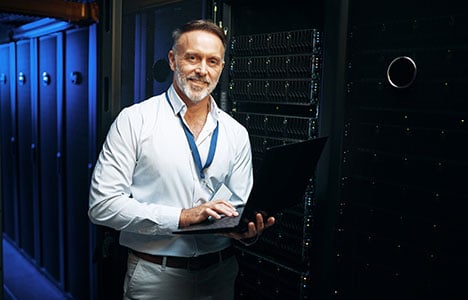 Man in a white shirt and blue neck lanyard holding a laptop in a server room